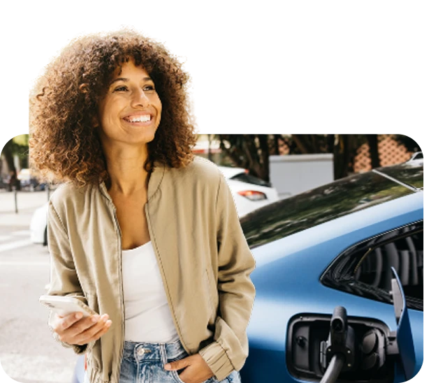 Smiling woman with curly hair holding a phone beside a blue electric car at a charging station.