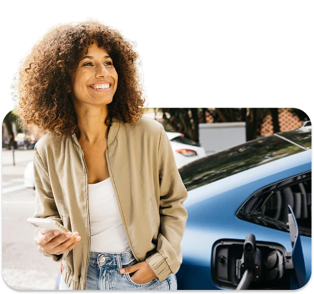 Smiling woman with curly hair in a beige jacket stands beside a blue car, holding a smartphone.