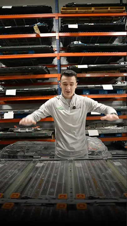 Warehouse worker in a gray polo shirt at a conveyor belt with tall orange racks of parts behind him.