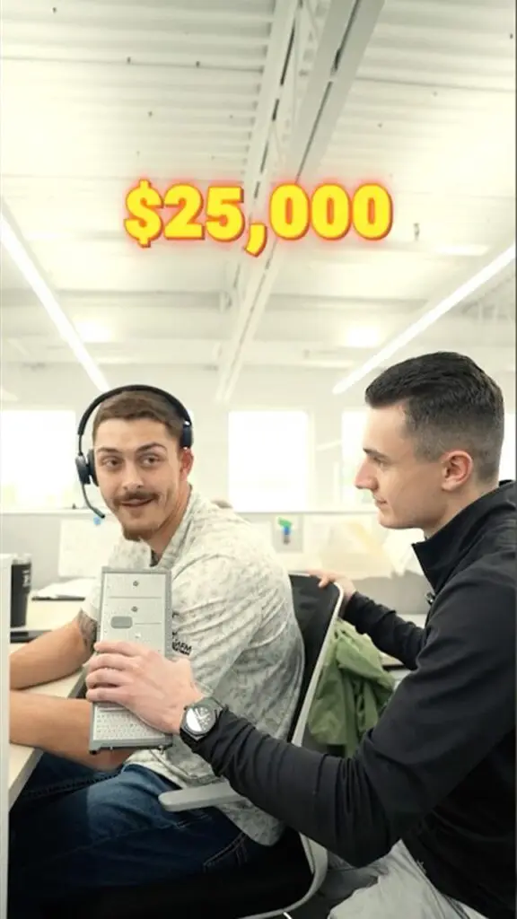 Two coworkers in a bright open-office; one wearing a headset holds a large computer component (likely a motherboard) while the other looks on, with a bold '$25,000' graphic above.