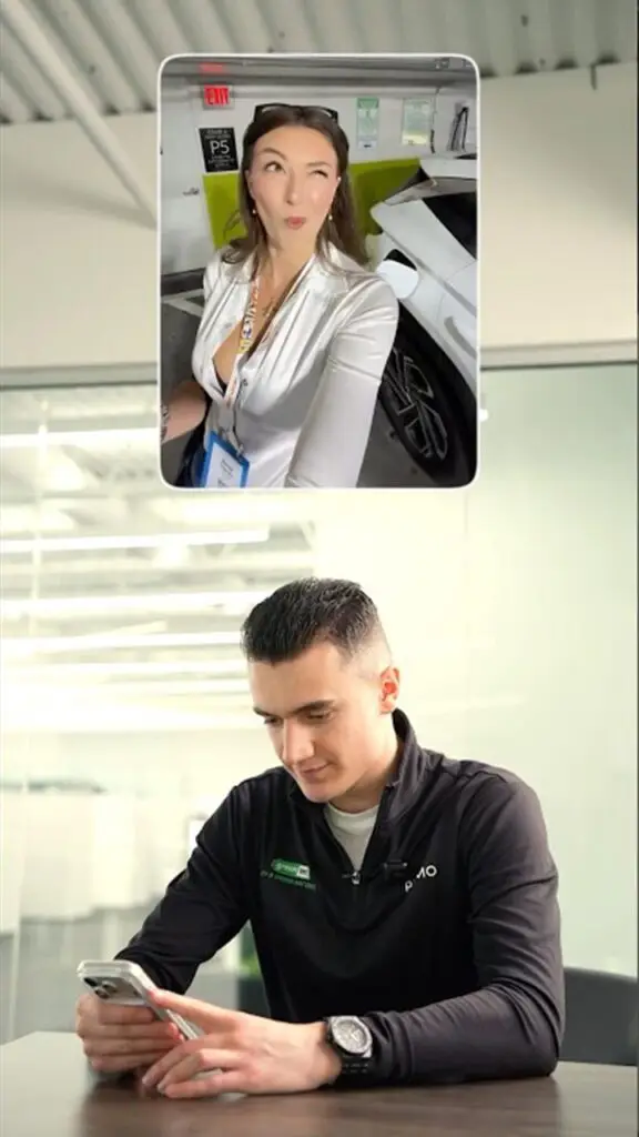Young man in a dark polo looks at his phone during a video call; a floating screen shows a woman in a white blouse posing in a parking garage