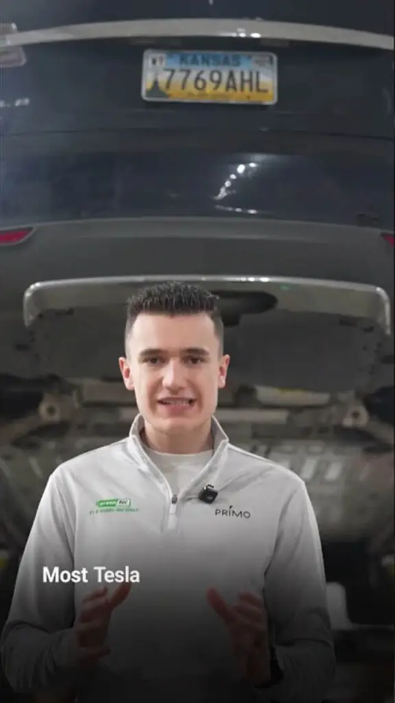 Presenter in a gray Primo shirt explains something in a car service bay with a raised vehicle behind him, captioned 'Most Tesla' on screen.