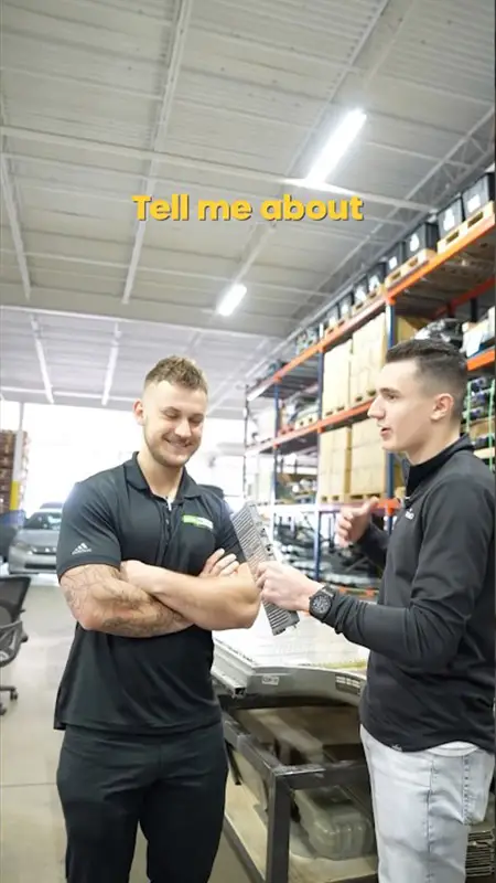 Two coworkers in a warehouse discuss documents at a metal worktable with industrial shelving in the background.