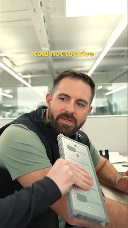 Man in a green shirt sits at a desk in an office, holding a large rectangular metal part with a concerned expression; the caption 'told not to drive' appears in yellow above.