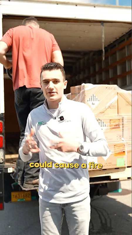 Man in a white pullover facing the camera, gesturing, in front of an open truck loaded with crates; caption reads 'could cause a fire'.
